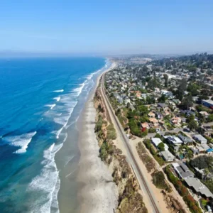 Del Mar Aerial view of the Del Mar coastline in San Diego with blue waves, sandy beach, a rail line, and coastal homes along the bluff.